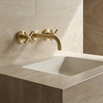 Brushed Gold cross handle taps with wall spout on a beige tiled wall with a white sink.