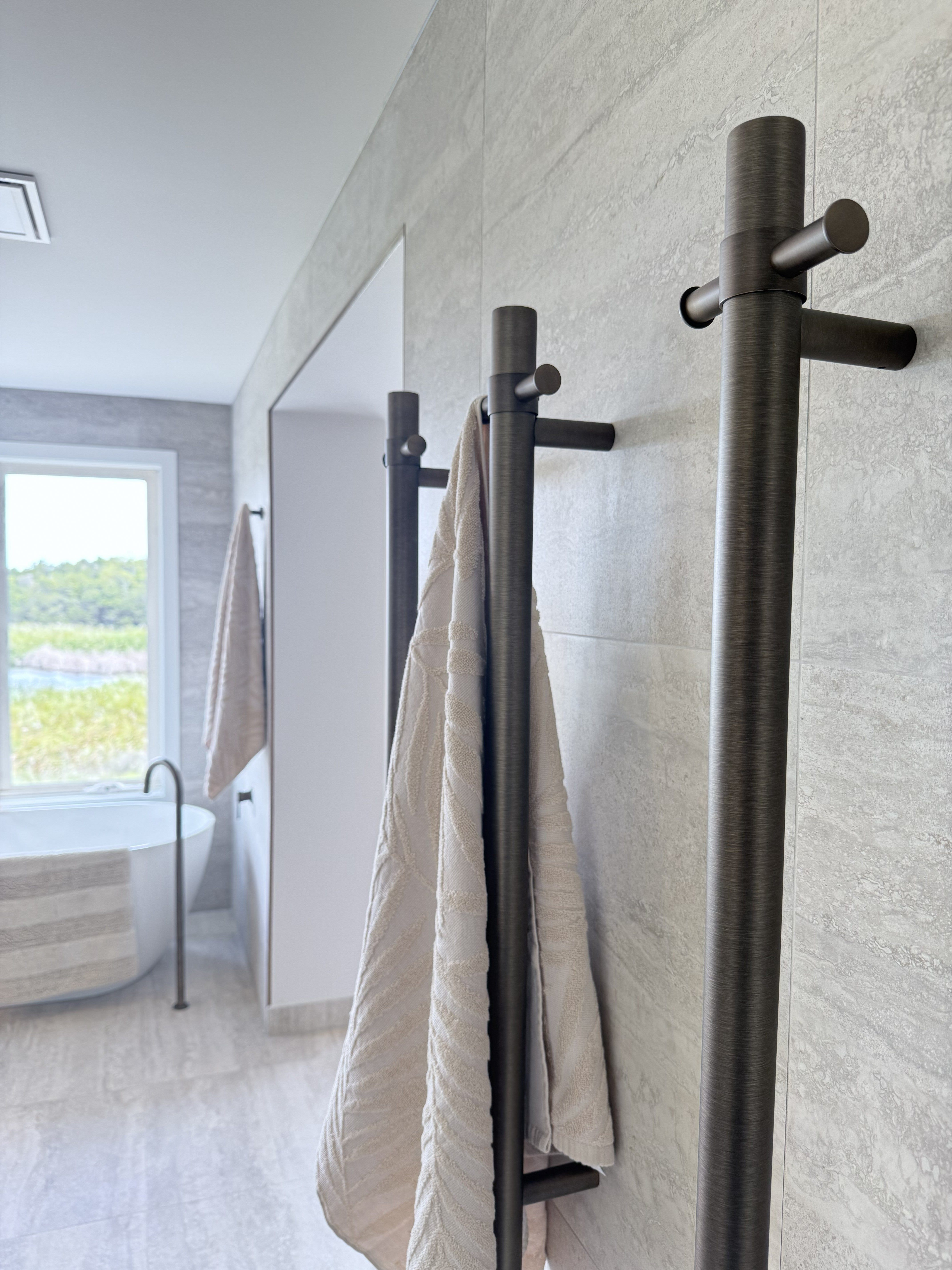 Bathroom with antique brass single bar heated towel rails and beige towels on a light grey tiled wall.