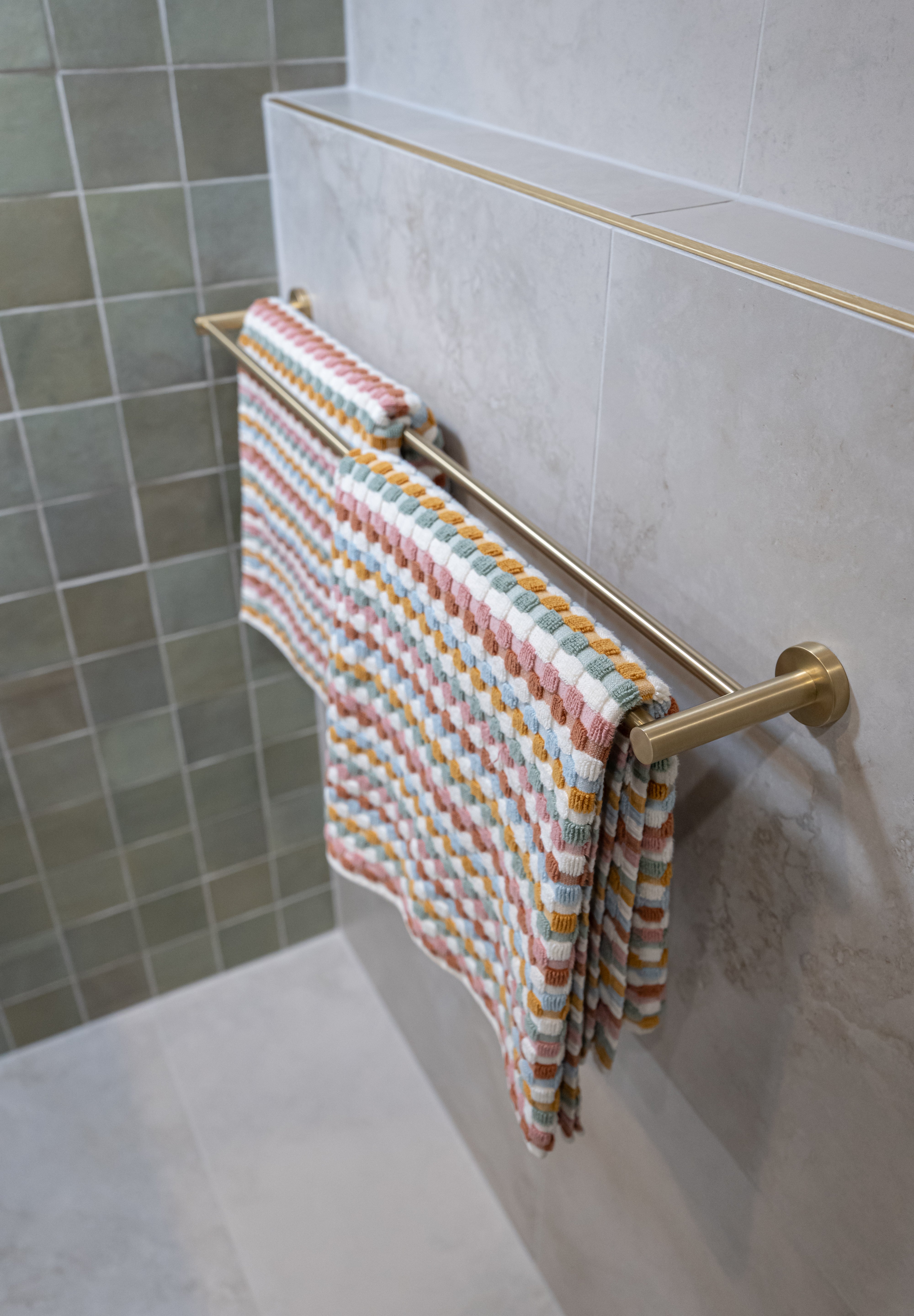 Brushed gold towel rail with a colorful striped towel against a tiled bathroom wall.