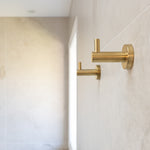 two brushed gold robe hooks mounted on a tiled wall