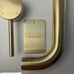 Brushed gold bathroom fixtures including a sample disc, wall mixer and wall spout  on a grey background.