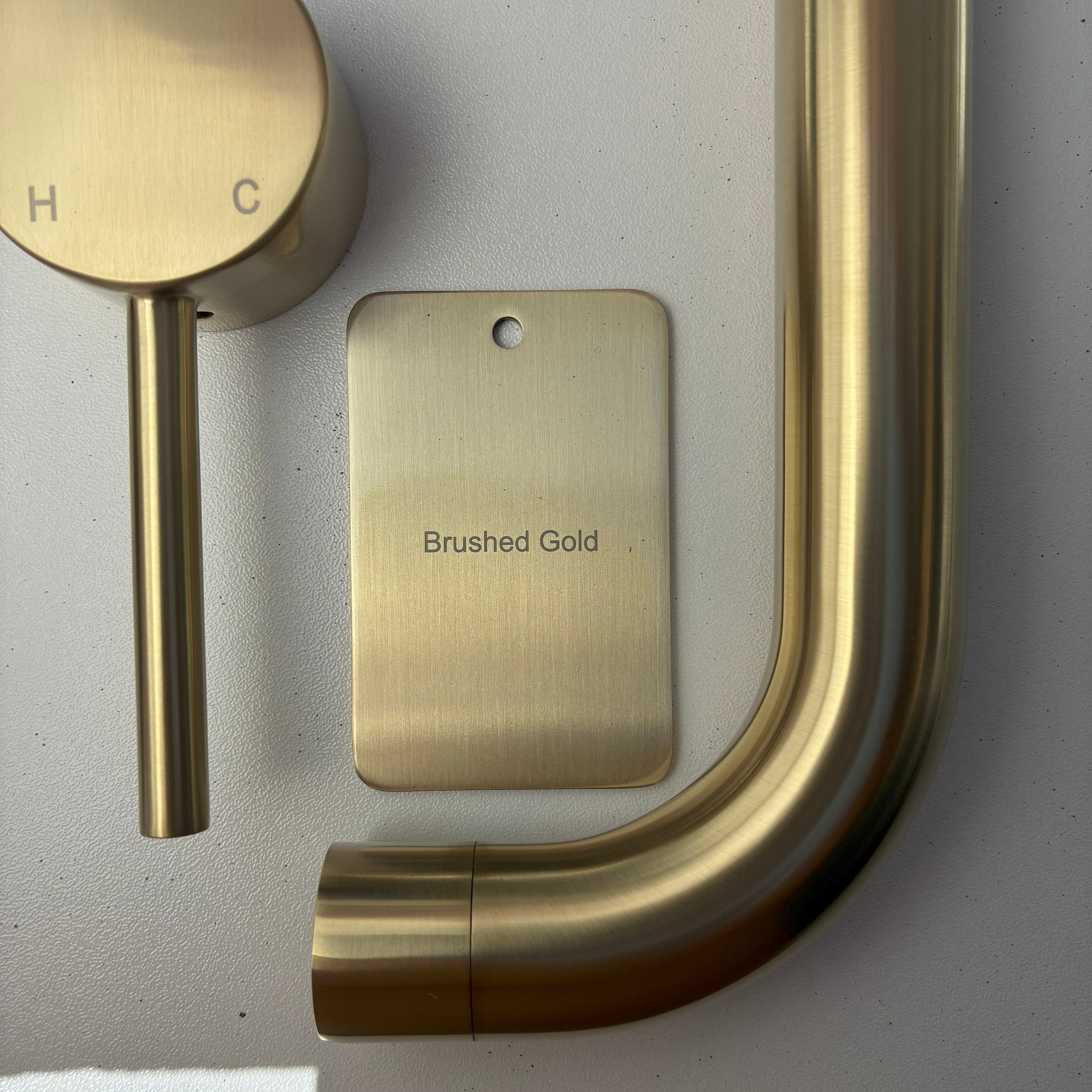 Brushed gold bathroom fixtures including a sample disc, wall mixer and wall spout  on a grey background.