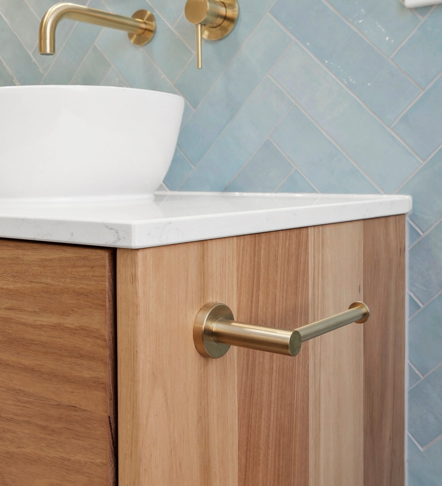 brushed gold toilet roll holder mounted on a white stone and timber vanity with blue tiles in the background