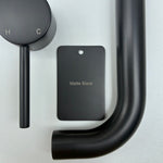 Matt black bathroom fixtures including a sample disc, wall mixer and wall spout  on a grey background.